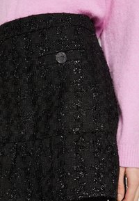 Gros plan sur une personne portant une jupe noire texturée avec une poche à bouton et un pull à manches longues rose pâle, la main détendue sur le côté.