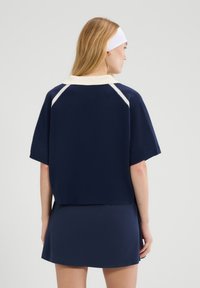 Kobieta ubrana w granatową koszulkę polo z krótkim rękawem, białym kołnierzykiem i paskami, granatową spódnicę oraz białą opaskę na głowę, zwrócona tyłem na jednolitym tle.