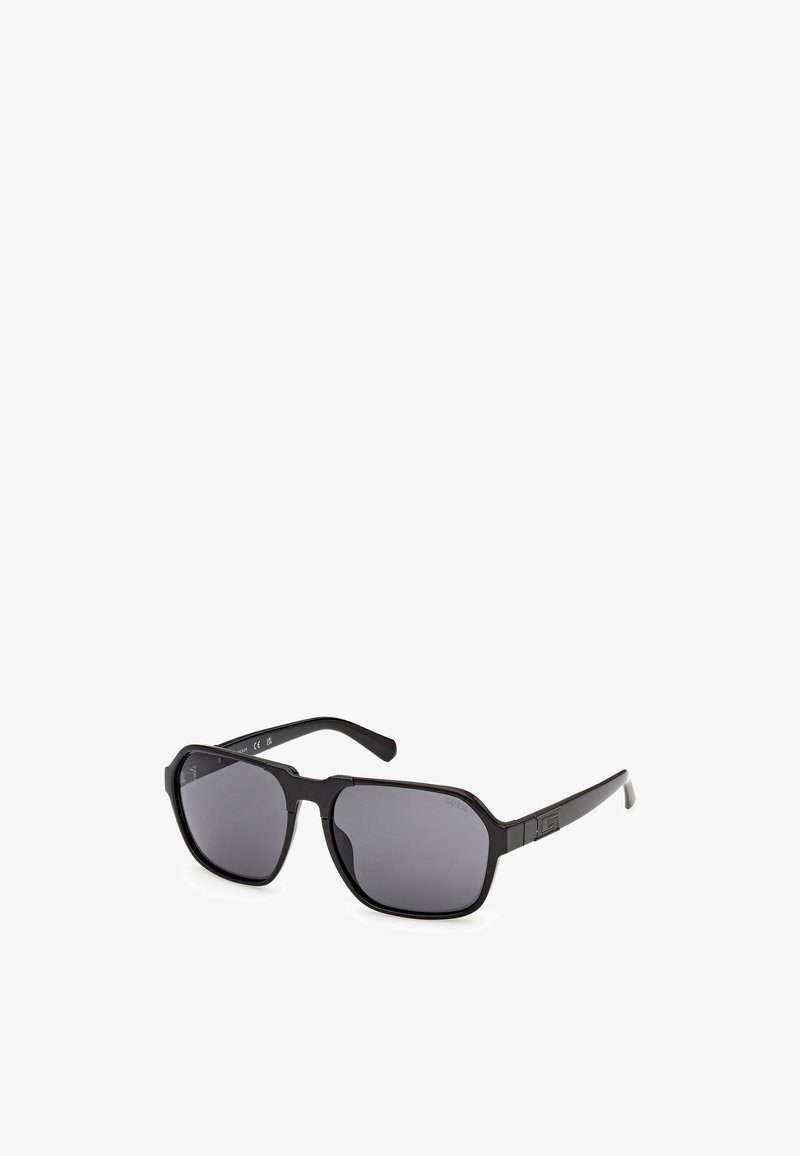 Lunettes de soleil rectangulaires noires avec des verres foncés et un marquage subtil sur les branches, présentées sous un léger angle sur un fond blanc.
