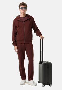 Bordeaux rits hoodie en bijpassende broek, witte sneakers; zwarte harde koffer met vier wielen en uitschuifbare handgreep.