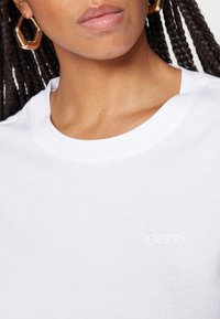 Gros plan d'une personne portant des boucles d'oreilles créoles hexagonales en or et une chemise blanche avec « BOSS » brodé sur la poitrine.