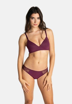 Femme aux longs cheveux foncés portant un soutien-gorge bordeaux assorti et une culotte bordée de dentelle, debout devant un fond blanc uni.