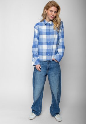 Frau mit langen blonden Haaren, die ein blau-weiß kariertes Hemd mit Knöpfen, lockere blaue Jeans und weiße Sneakers trägt, steht vor einem einfarbigen Hintergrund.