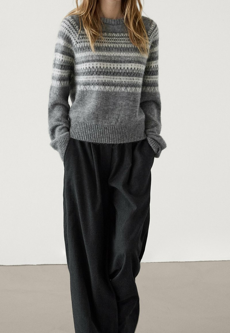 Femme portant un pull en maille gris à motifs, les mains dans les poches, et un pantalon noir ample, sur un fond uni.