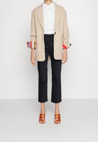 Beige oversized kavaj med flerfärgade randiga ärmar, vit skjorta med färgglada knappar, svarta korta byxor och orange remsandaler.