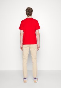 Rode T-shirt met korte mouwen van katoen, beige broek en witte sneakers met blauwe en rode accenten, gezien van achteren.