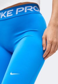 Κοντινό πλάνο ενός ατόμου που φοράει λαμπερά μπλε συμπιεστικό κολάν Nike Pro με λευκό λογότυπο Nike και μερικό χέρι που ακουμπάει στον μηρό.