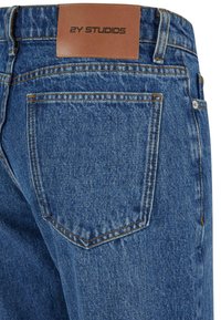 Denimske hlače v modrem pranju, z usnjenim oznako na pasu z napisom "2Y STUDIOS" in vidnim šivom na zadnjih žepih.