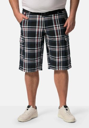 Mand iført sorte, hvide og røde ternede cargoshorts med lommer, hvide sneakers og en hvid skjorte, stående med hænderne i lommerne.