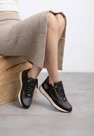 Las zapatillas negras y grises presentan un acabado texturizado, herrajes dorados y una cremallera lateral. La suela es blanca con un acento en marrón claro.