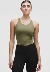 Una donna che indossa un top aderente senza maniche verde oliva e leggings neri a vita alta, in piedi contro uno sfondo chiaro e semplice.