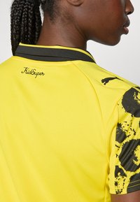 Osoba s vrkôčkami vo vlasoch, oblečená v športovom tričku žlto-čiernej farby s nápisom "KidSuper" a logom Puma na ramene a golieri.