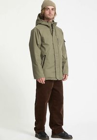 Olijfgroene waterdichte jas met capuchon, ritssluiting aan de voorkant en ritssluitingszakken; gecombineerd met bruine corduroy broek en zwarte schoenen.