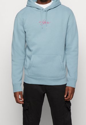 Mann trägt eine hellblaue Kapuzenjacke mit Fronttasche und pinkem Schriftzug, kombiniert mit schwarzen Cargohosen.