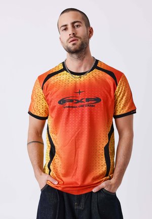 Uomo con capelli corti e barba, indossa una maglietta sportiva arancione e gialla con dettagli neri e logo, mani nelle tasche, sfondo neutro.