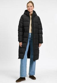 Lächelnde Frau, die einen langen schwarzen Puffer-Mantel mit Kapuze, einen hellbeigen Pullover, blaue Jeans und braune Sneaker trägt, vor einem weißen Hintergrund steht.