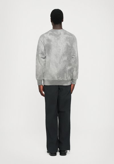 Ein Mann, der mit dem Rücken zur Kamera steht, trägt einen hellgrauen Batik-Sweater, schwarze weit geschnittene Hosen und schwarze Schuhe vor einem schlichten weißen Hintergrund.