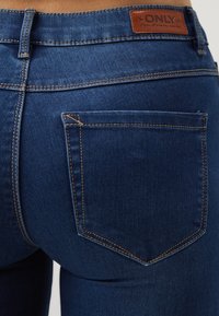 Jeans de denim azul oscuro con corte recto, que cuentan con un bolsillo trasero, costuras naranjas y una etiqueta de marca de cuero marrón en la cintura.
