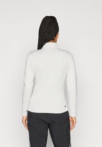 Top blanco de manga larga con cuello alto y una textura suave. Presenta un pequeño logo en el dobladillo y se combina con pantalones oscuros y texturizados.