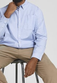 Camicia azzurra chiara con taschino sul petto e maniche lunghe, abbinata a pantaloni color khaki. Il tessuto sembra liscio e leggero.