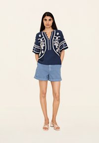 Marinblus med broderade detaljer i vitt, V-ringning och korta ärmar, samt ljusblå rullade shorts och sandaler.