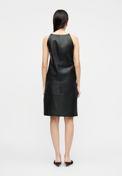 Γυναίκα με αμάνικο μαύρο δερμάτινο φόρεμα και μαύρα τακούνια mule, στέκεται με την πλάτη σε απλό ανοιχτό γκρι φόντο.