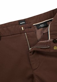 Bruna slim-fit byxor med dragkedja, metallknapp och en slät tygkänsla med en subtil lyster. Etiketter synliga invändigt i linningen.