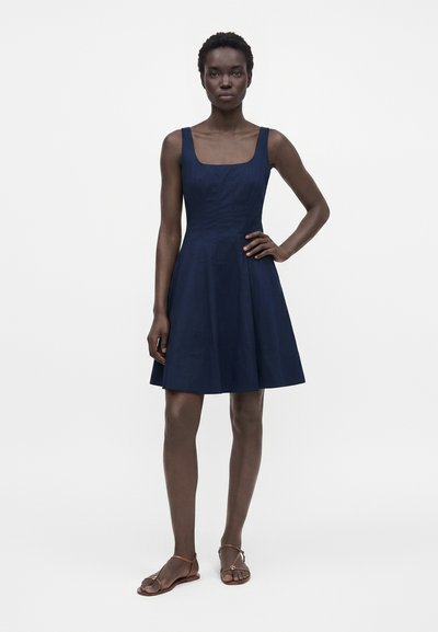 Femme debout portant une robe bleu marine sans manches, longueur genou, avec un décolleté carré et des sandales plates marron, une main sur la hanche.