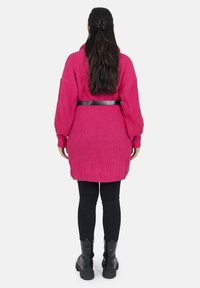 Rosa stickad cardigan med ribbad textur, långa ärmar och ett svart bälte i midjan. Buren över svarta leggings och kraftiga svarta stövlar.