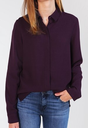 Person iført en mørklilla skjorte med lange ærmer og blå jeans, med den ene hånd delvist i lommen.