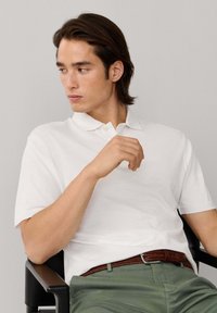 Jeune homme aux cheveux bruns mi-longs, portant un polo blanc et un pantalon vert, assis sur une chaise noire regardant sur le côté.