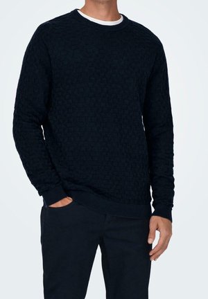 Mand iført tekstureret sort langærmet sweater over hvid undertrøje med mørke bukser, den ene hånd i lommen og den anden afslappet langs siden.