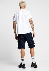 White short-sleeve T-shirt, navy cargo shorts with pockets, and black sneakers. Model has light hair and visible tattoos on arms.