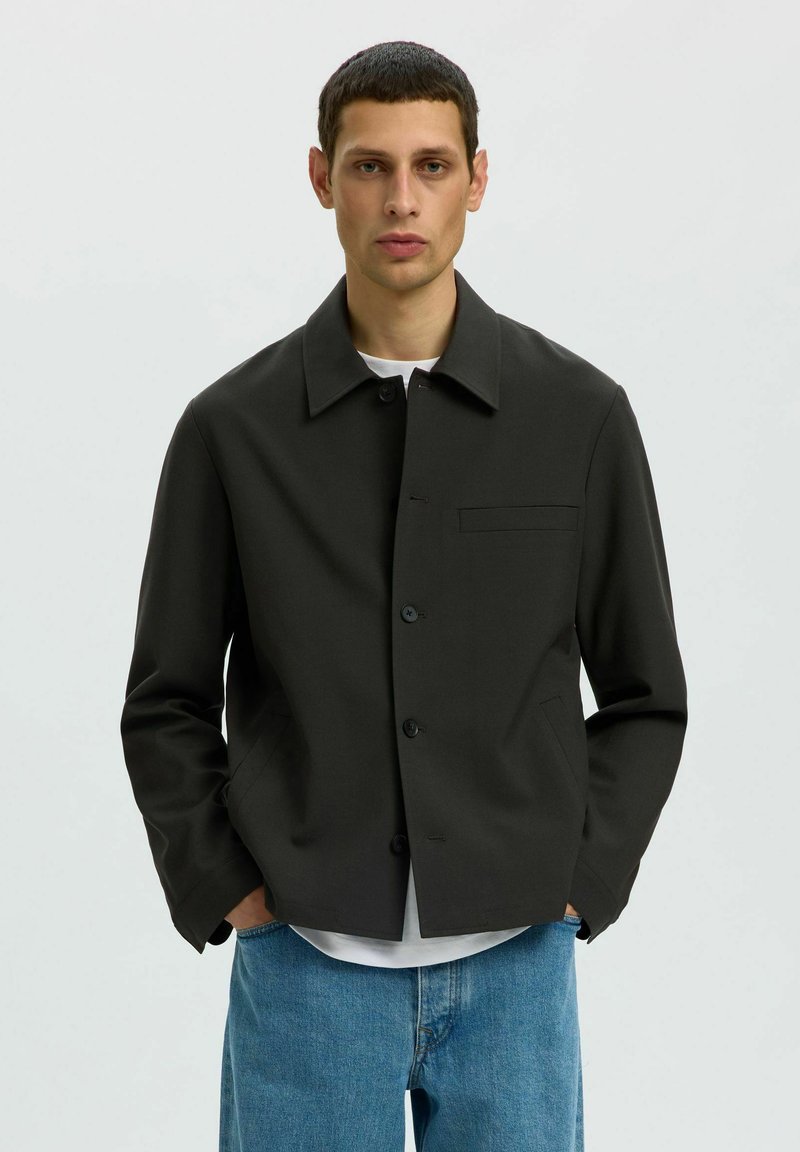 Homme portant une veste noire boutonnée sur une chemise blanche avec un jean bleu, debout les mains dans les poches devant un fond uni.