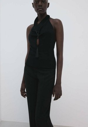 Femme portant un haut noir sans manches style halter et un pantalon noir, accessoirisée d'un long collier avec un pendentif à pompon.