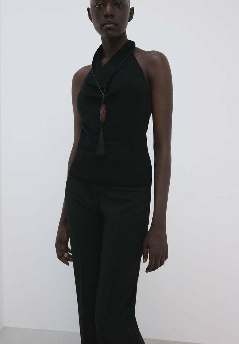 Femme portant un haut noir sans manches style halter et un pantalon noir, accessoirisée d'un long collier avec un pendentif à pompon.