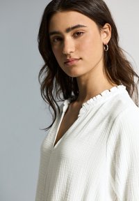 Blouse blanche texturée avec un col à volants et une découpe en V. Fabriquée en tissu léger et doux, idéale pour une tenue décontractée.