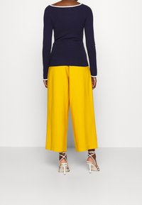 Osoba stojící, oblečená v námořnickém dlouhém rukávu, jasně žlutých kalhotách s širokými nohavicemi a bílých sandálech na vysokém podpatku s pásky kolem kotníků.