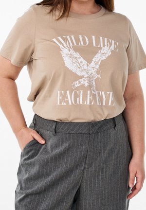 Personne portant un t-shirt beige avec un aigle blanc et le texte "WILD LIFE EAGLE EYE", associé à un pantalon gris à fines rayures.