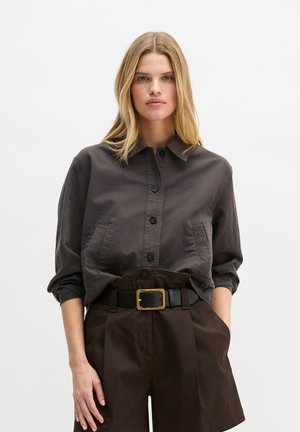 Kobieta ubrana w ciemną koszulę na guziki z podwiniętymi rękawami, wpuszczoną w brązowe szorty z wysokim stanem, z czarnym paskiem i złotą klamrą.