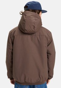 Person som bär en brun huva-jacka och blå keps, vänder bort ansiktet, står mot en plain vit bakgrund.