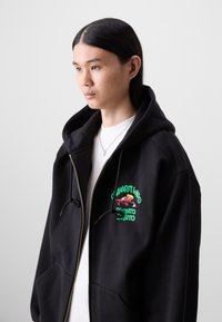 Black zip-up hoodie with a drawstring hood, featuring a colorful graphic on the chest. It has a relaxed fit and front pockets.