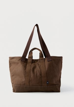 Grote bruine canvas draagtas met zowel korte als lange hengsels, drukknoopsluiting en een klein blauw stoffen label onderaan.