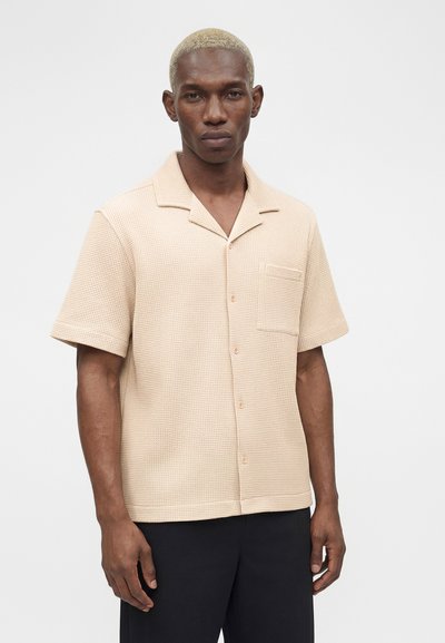 Άνδρας με κοντά ξανθά μαλλιά φοράει μπεζ υφασμάτινο κοντομάνικο πουκάμισο με κουμπιά και μαύρο παντελόνι πάνω σε απλό λευκό φόντο.