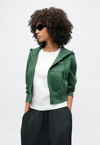 Zelená mikina na zips s dvoma prednými vreckami a biela tričko s okrúhlym výstrihom pod ňou, doplnené o čierne skladané nohavice.