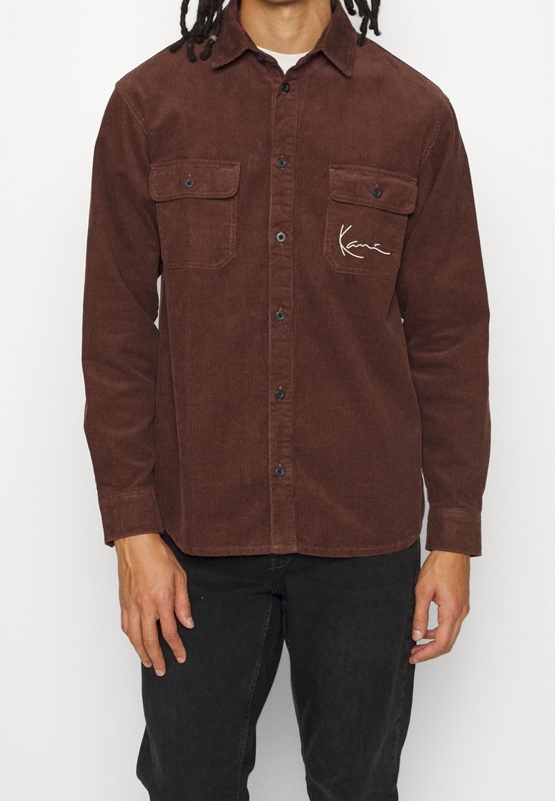 Braunes Cordhemd mit einer Knopfleiste, zwei Brusttaschen und einem signifikanten Logo auf der linken Seite. Es hat lange Ärmel und einen Kragen.