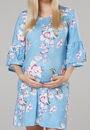Kobieta w ciąży z długimi blond włosami, ubrana w niebieską kwiatową sukienkę, trzymająca brzuch obiema rękami na neutralnym tle.