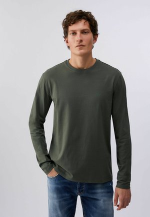 Jeune homme aux cheveux courts et bouclés portant une chemise à manches longues vert olive et un jean bleu, debout avec une main dans la poche.