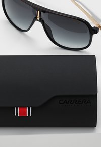 Ochelari de soare cu ramă neagră și lentile gri cu degradé, alături de o etui mată neagră cu un accent în dungi roșii și albe și logo.