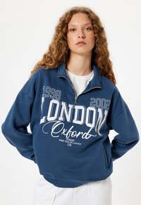 Sudadera de color azul oscuro con un alto cuello, cierre de cremallera, texto gráfico blanco que presenta "LONDRES" y "Oxford", y puños y dobladillo de canalé.